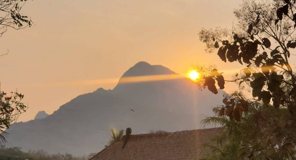 Sunrise breaking over a hazy mountain ridge, seen through trees above a thatched roof