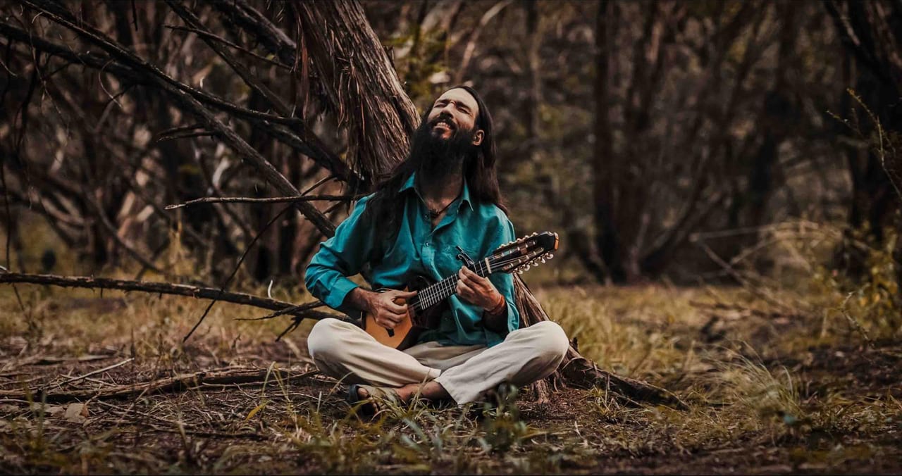 Michael playing charango, seated on the ground among trees